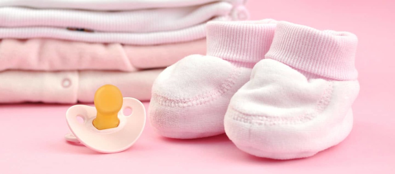 A pile of folded pink baby clothes, a pair of pink booties, and a dummy.