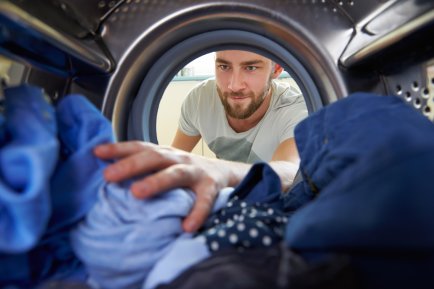 A bearded man reaching into a washing machine drum full of blue clothing.